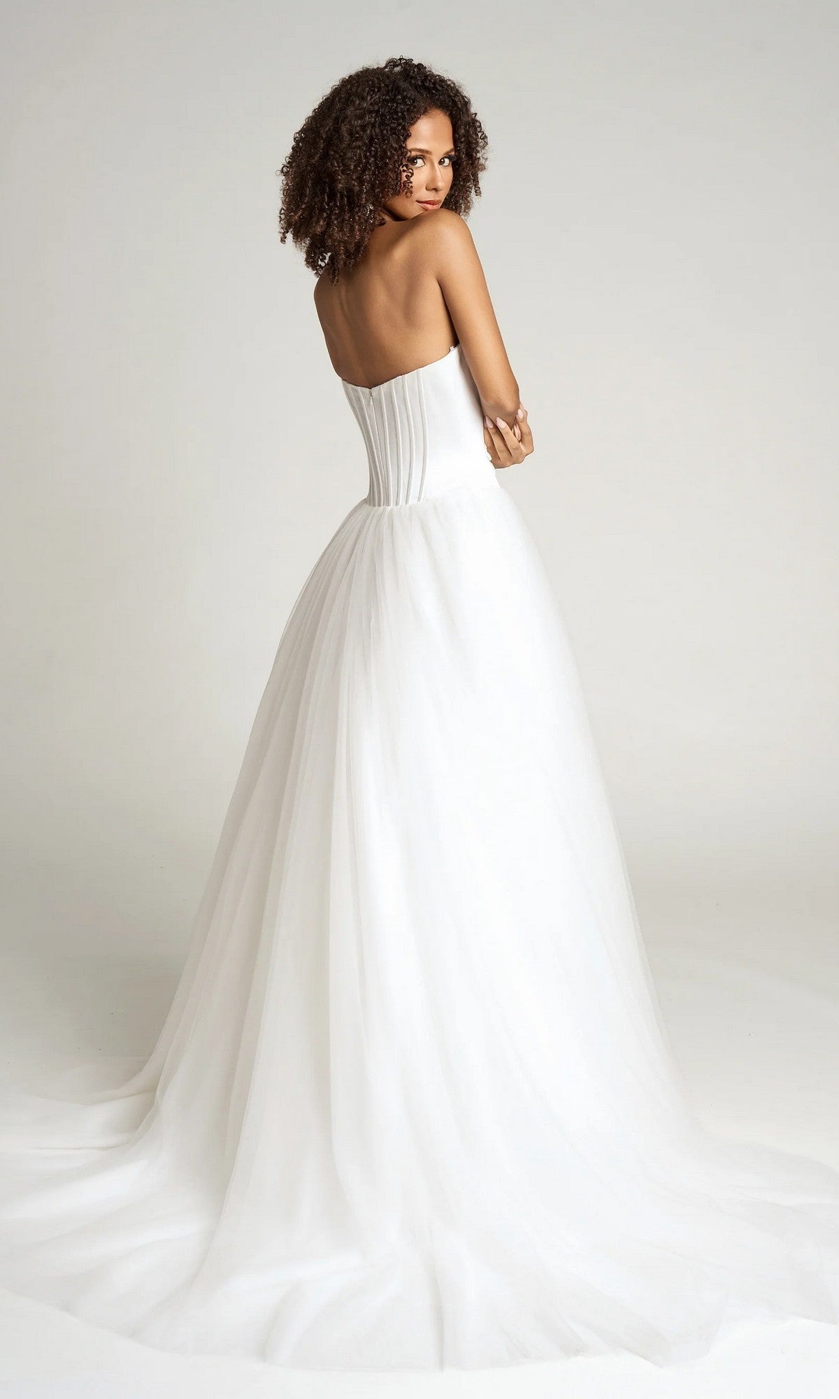 Woman wearing the Portia and Scarlett PS26102 strapless ball gown, featuring a corset bodice and full tulle skirt, stands looking over her shoulder against a plain background.